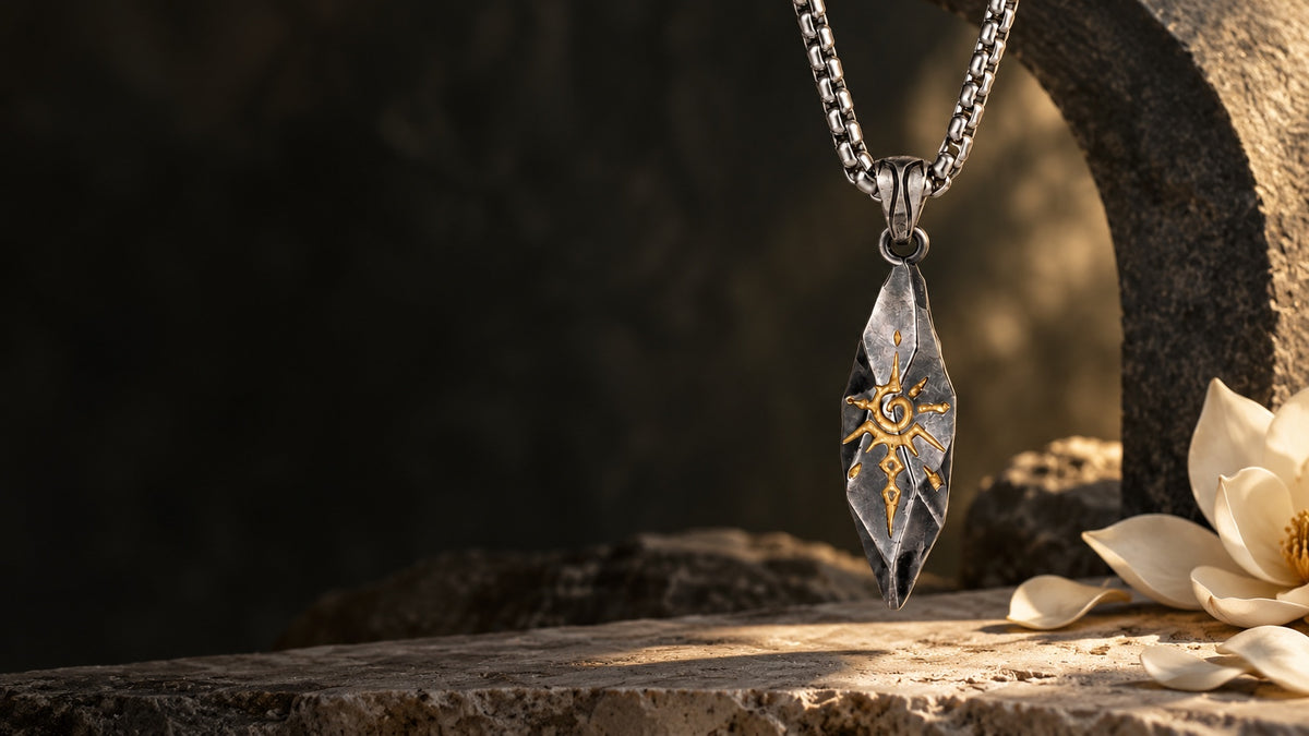 Sigil Graphite necklace with its gold sun motif, styled beside charcoal stone and ivory magnolia.